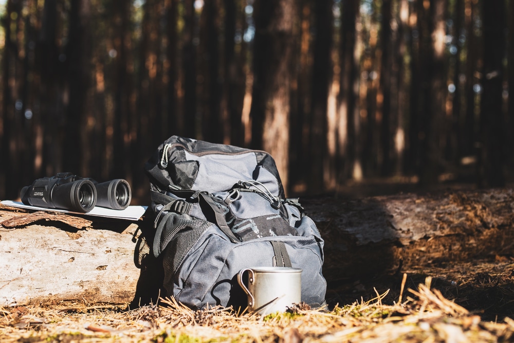 Grey hiking backpack resting against a fallen log next to binoculars and a metal camp mug