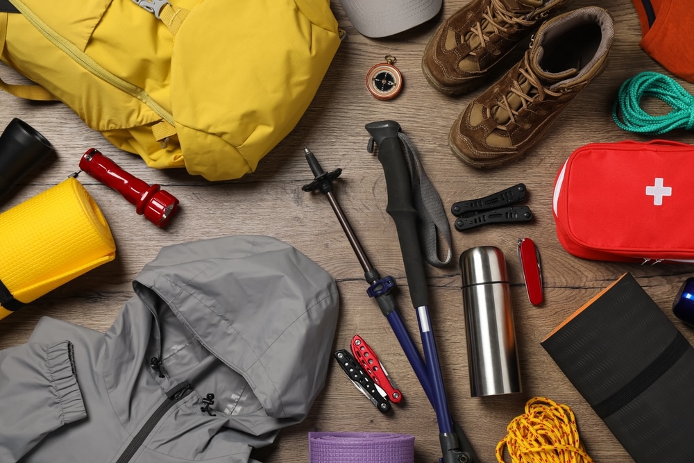 Flat lay of hiking and camping gear including a yellow backpack, boots, jacket, and first aid kit on a wooden surface