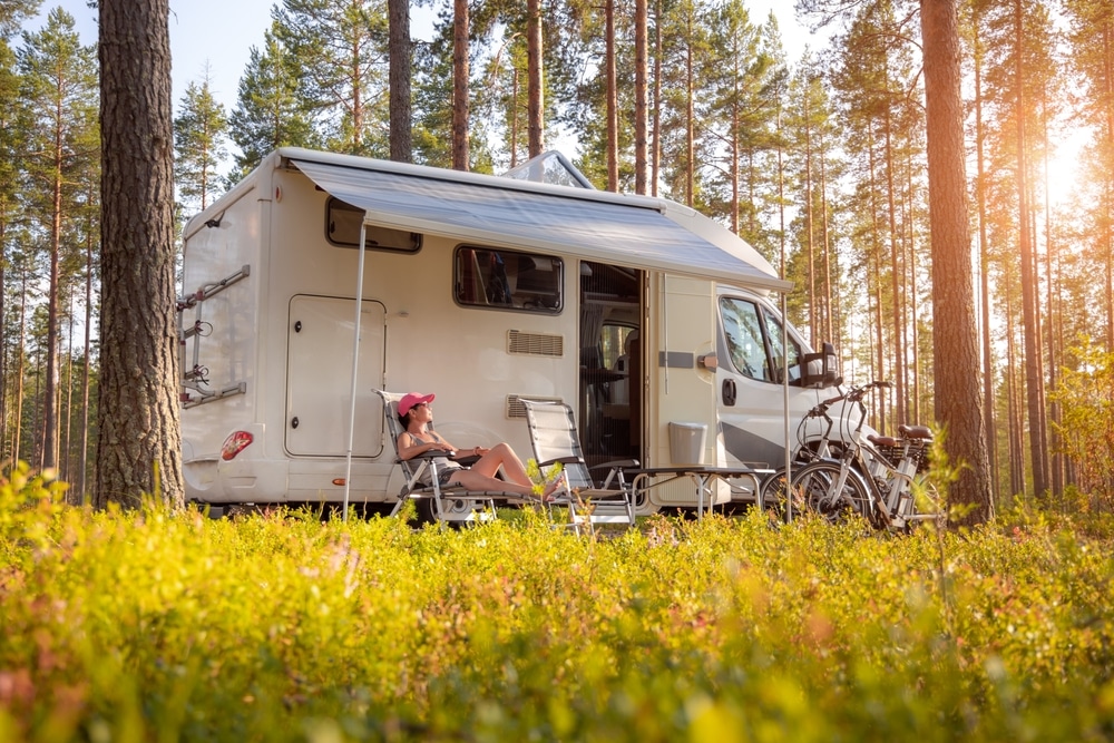 Woman relaxing in a lawn chair next to a white motorhome parked in a sunny pine forest
