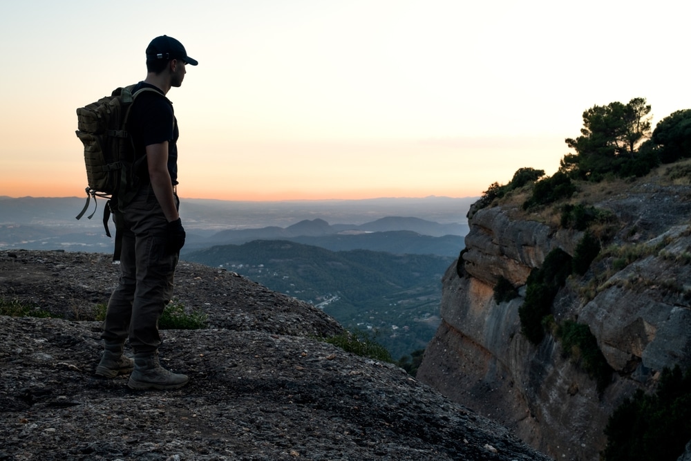 Hiker with a tactical backpack standing on a rocky cliff edge looking at a mountain valley at sunset