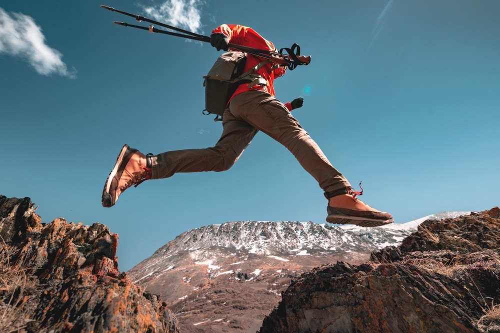 Low angle shot of a hiker in an orange jacket jumping over a rocky gap in the mountains