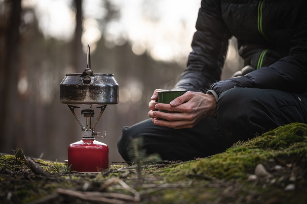 Camper sitting on mossy ground in a forest holding a mug next to a portable camping stove