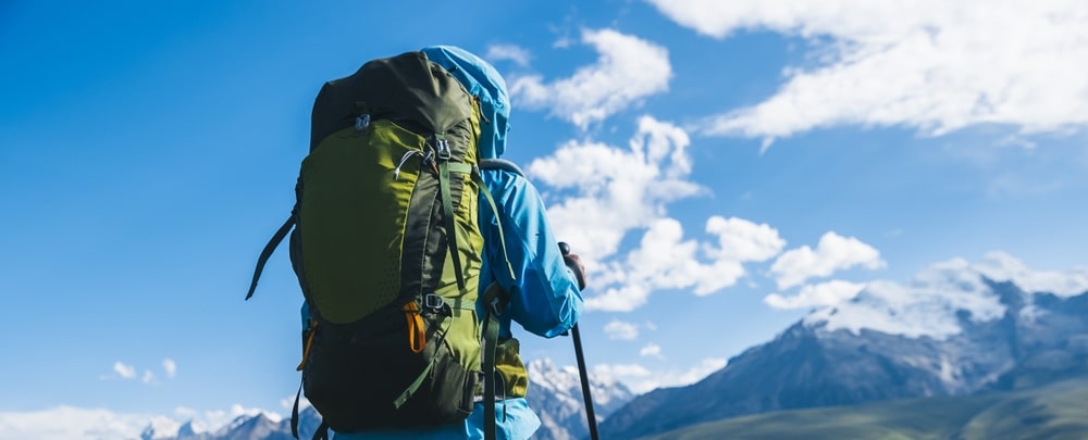 Hiker with a large green backpack looking at snowy mountains under a blue sky