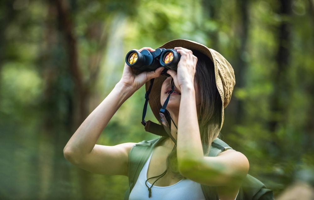 Woman wearing a hat looking through binoculars in a dense forest